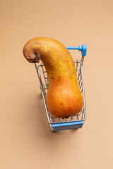 Wall Mural - One ugly curved pear in small blue supermarket cart on beige background. Vertical orientation.