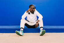 Young Afro American Black Man Wearing A White Sweatshirt, Basketball Sneakers, Sunglasses, Comb In The Hair And Headphones Seated On A Blue Wall Looking To The Side With Confidence And Attitude