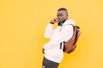 Wall Mural - Young african American black man against a yellow wall wearing a white sweatshirt and a backpack listening music on wireless headphones while looking at camera with attitude