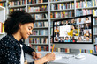 © Kateryna - A smiling African American female student takes notes during lecture, listens and watches online lesson while sitting in a library. Side view at a girl and computer with different multinational people