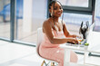 Fashionable photo of young beautiful romantic african american woman with glass of wine, wearing pink dress working with laptop.