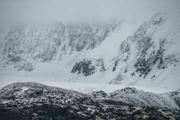 Foggy mountain landscape with white snow on black rocks in cloudy weather. Misty mountain minimalism of snowbound mountain in low clouds. Minimalist nature background of snowy mountainside in fog.