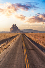 View Of A Road In A Dry Desert With A Shiprock Mountain Peak In The Background. Sunrise Sky Art Render. Rattlesnake, New Mexico, United States. Concept: Road Trip, Adventure, Travel