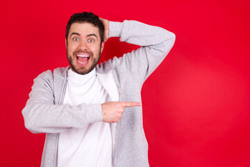 Wall Mural - young handsome caucasian man in sports clothes against red wall feeling positive has amazed expression, indicates something. One hand on head and pointing with other hand.