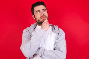 Portrait of thoughtful young handsome caucasian man in sports clothes against red wall keeps hand under chin, looks away trying to remember something or listens something with interest. Youth concept.