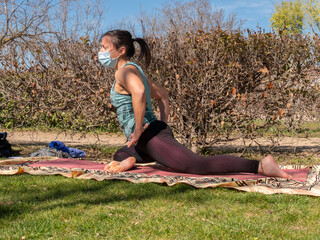 A brunette woman doing yoga class in a park on a sunny day and wearing a face mask