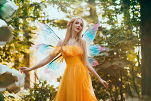 Portrait Fantasy Woman Blonde Forest Fairy. Elf Girl Fashion Model In Bright Yellow Dress, Butterfly Wings. Pixie Walks In Summer Nature. Green Spring Tree, Wood, Sun Light Magic Radiance. Long Hair