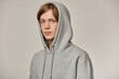 Cool looking male, handsome guy with blond hair. Wearing grey hoodie and puts hood on. People and emotion concept. Watching serious at the camera isolated over grey background