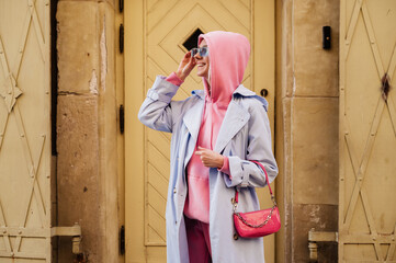 Wall Mural - Happy smiling fashionable woman wearing trendy sport chic style outfit posing in street of European city. Blue trench coat, sunglasses, pink hoodie, mini baguette bag. Copy, empty space for text