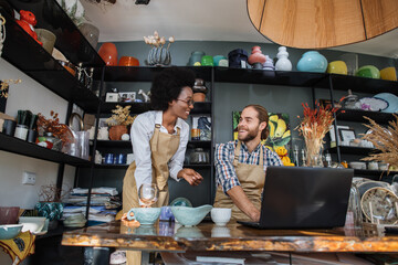 Wall Mural - Smiling african woman and caucasian man working together st decor shop and using laptop. Two colleagues doing inventory of goods with help of modern gadget.