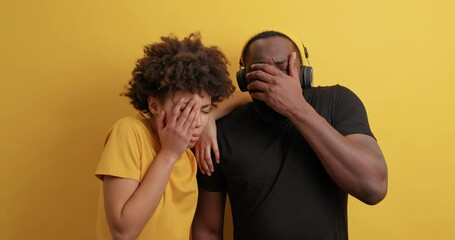 Wall Mural - Dark skinned woman and man concentrated surprisingly at camera then cover faces not to see something horrible feel scared while watching horror film dressed in casual clothes isolated on yellow wall