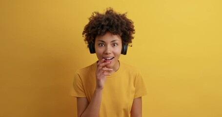 Wall Mural - Beautiful dark skinned woman looks with great wonder at camera says wow smiles broadly wears casual t shirt wireless headphones on ears hears impressive news isolated over yellow background.