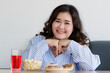 © Bangkok Click Studio - Closeup shot of Asian young happy beautiful friendly overweight fat chubby female sit relax smiling look at camera behind table with red sweet water popcorn donut and cake in blurred foreground