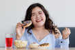 © Bangkok Click Studio - Happiness closeup shot of Asian young happy beautiful hungry overweight fat chubby female smile at camera hold chocolate donut in hand and hold fist up satisfy with dessert cake popcorn on table