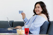 © Bangkok Click Studio - Asian young happy beautiful fat chubby female shopper sit relaxing on sofa smiling at camera hold blue credit card on hand while using laptop computer buy products shopping and make payment online