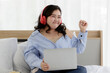 © Bangkok Click Studio - Asian young happy funny fat chubby oversized woman wears big red headphones around head listening to music and sit enjoy   alone on bed using laptop computer play playlist at home in bedroom