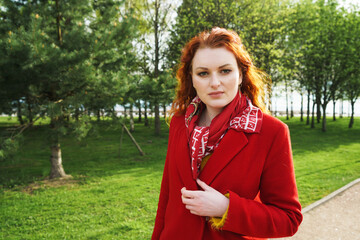 Portrait of cute caucasian red-haired woman in a red coat against the background of a green spring park