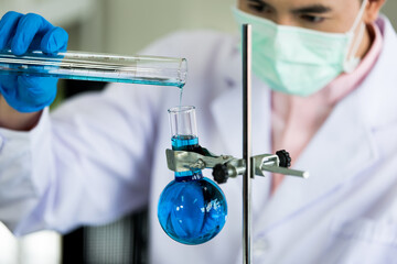 scientific male researcher in white coat, green mask, and blue gloves pouring blue liquid specimen i