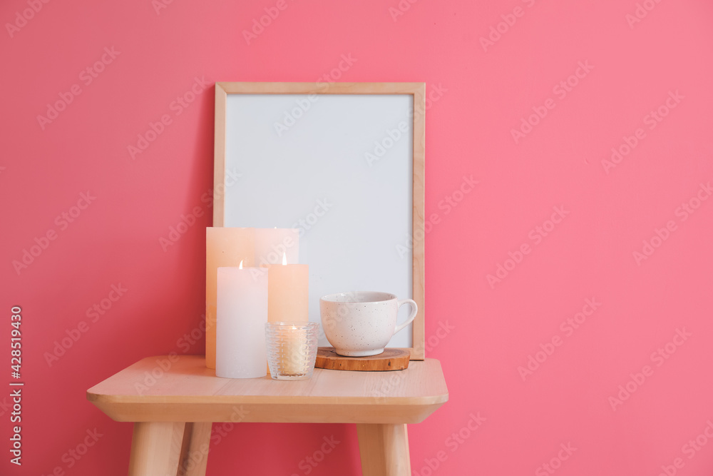 Burning candles, cup and blank frame on table against color background