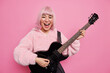 Positive fashionable female rock star with pink hairstyle plays acoustic guitar has own music band dressed in stylish coat creats new song for her album poses indoor. Happy stylish woman guitarist
