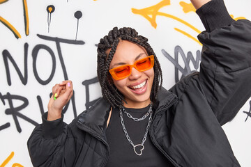 Wall Mural - Energetic pleased dark skinned woman with braids expresses positive emotions dances carefree wears orange shades fashionable jacket poses against graffiti wall in public place. Teenagers lifestyle