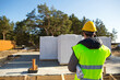 © Ольга Симонова - The builder  talking on the phone. Architect in a protective yellow helmet and a signal vest stands backs in front of the construction site of a house with a foundation and aerated concrete blocks.