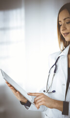 Wall Mural - Young woman-doctor is holding a tablet computer in her hands, while standing in a clinic. Portrait of friendly female physician with a stethoscope at work. Perfect medical service in a hospital