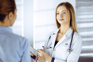 Wall Mural - Young woman-doctor and her patient are discussing patient's current health examination, while standing together in a hospital office. Female physician is writing some marks, using a clipboard. Perfect