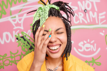 Wall Mural - Stylish hipster girl has dreadlocks smiles broadly shows golden teeth makes face palm being very happy stands against large graffiti wall expresses happiness. Youth lifestyle emotions urban life