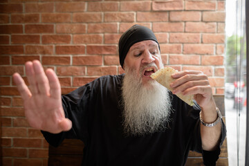 Hungry man with long gray beard eating taco while getting disturbed and showing stop gesture