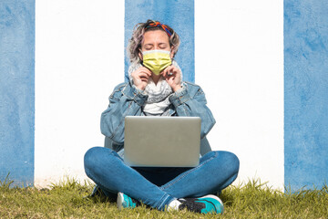 Poster - Spanish woman in a face mask using her laptop against a blue and white wall in the park