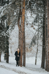 Poster - Handsome young man in a black winter outfit with a brown scarf leaning against a forest tree trunk