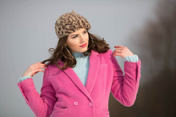 Wall Mural - Young brunette girl standing in the snow wearing a pink coat with a stylish hat and big earing