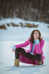 Wall Mural - Young thoughtful brunette girl with eyeglasses  kneeling down in the snow forest wearing a pink coat