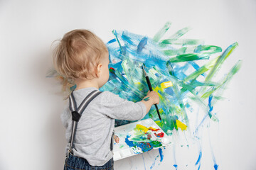 Little child with a brush and a palette of paints in his hands draws on a white wall. Happy childhood