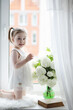 © alexkich - A little girl is sitting on the windowsill. A bouquet of flowers in a vase by the window and a girl sniffing flowers. A little princess in a white dress with a bouquet of white flowers by the window.