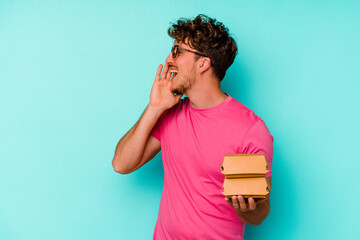 Young caucasian man holding two burgers isolated on blue background shouting and holding palm near opened mouth.