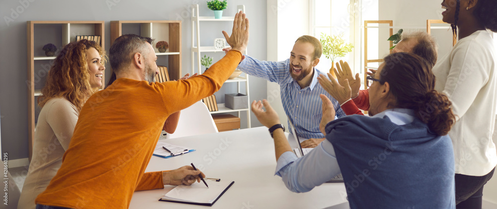 Happy successful multiracial business team clapping hands while two colleagues giving high fives gesture each other. Businesspeople laugh and cheer sitting at table in office on briefing meeting - obrazy, fototapety, plakaty 