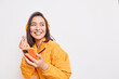 © wayhome.studio  - Studio shot of happy Asian millennial girl makes eastern like sign smiles gladfully looks away dressed in orange jacket uses smartphone and headphones for listening favorite music poses indoor