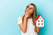 © Asier - Young caucasian woman holding a house model isolated on blue background looking sideways with doubtful and skeptical expression.