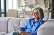 © insta_photos - Happy middle aged senior woman sitting on couch holding using digital tablet device during video call with family friends. Older female reading ebook or watching zoom video meeting online.