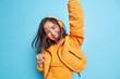 Joyful Asian girl has fun dark hair floaring in air while jumping keeps arms raised wears wireless headphones listens music feels energetic isolated over blue background. People lifestyle happiness