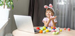 © Angelov - Pretty little girl wearing bunny ears and painting easter eggs at home during coronavirus covid-19 outbreak. Kid using laptop on kitchen, online, video call to friends