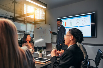 Team of young smiling business people in conference room discussing project. A speaker stands next to the flipchart and shows the data.