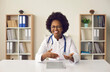 © Studio Romantic - Head shot portrait of smiling african american female doctor wearing eyeglasses, uniform with stethoscope speaking, consulting patient online, looking at camera, making video call sitting at table