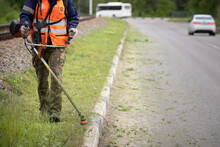 Closeup Of A Man With A Lawn Mower In His Hands. A Worker Mows The Grass On The Side Of The Road. Improvement Of The Adjacent Territory