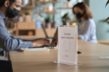 Closeup of social distance sign Keep Distance in focus standing on table. Blurred indian businessman and latin student working on computers in office university learning campus wearing face mask.