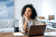 © Kateryna - A happy good looking successful curly African American stylish freelancer or student girl, sitting at her desktop at home office, looking to the side, smiling pleasantly, dreaming about vacation