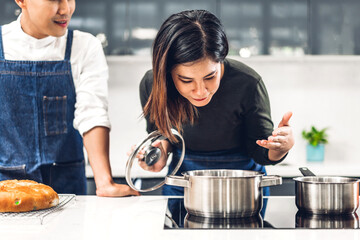 Wall Mural - Young asian family couple having fun standing near stove and cooking together.Happy couple looking and smelling tasting fresh delicious from soup in a pot with steam at white interior kitchen