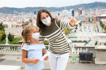 Wall Mural - Mom and daughter in protective masks walk along the street of a European summer city